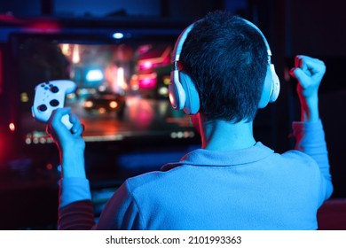 Boy In Headphones Plays A Video Game On The Big TV Screen. Gamer With A Joystick. Online Gaming With Friends, Win, Prize. Fun Entertainment. Teens Play Adventure Games. Back View. Neon Lighting