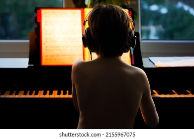 Boy In Headphones Sitting At The Piano In The Evening, View From The Back. Child Practicing Play Pianoat Home, Music Education Concept.