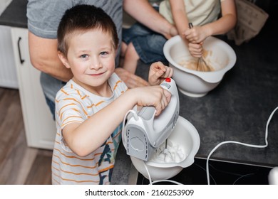 The boy helps to knead the dough with a mixer in a bowl in the kitchen at home. Satisfied child looks at the camera and smiles while cooking.