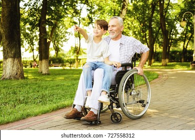 The boy and his grandfather are walking in the park. The old man is sitting on a wheelchair. The boy is sitting on the lap of an old man