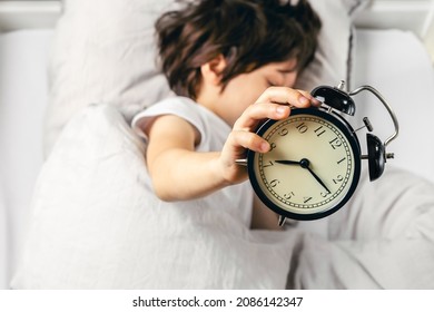 Boy is holding an alarm clock showing quarter past seven in the morning. Kid oversleeping and late for school.