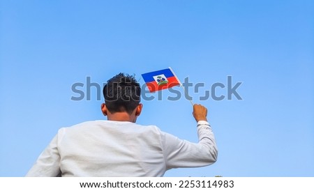 Boy holding Haiti flag against clear blue sky. Man hand waving Haitian flag view from back, copy space for text