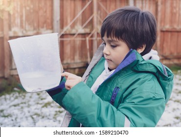 Boy Holding Measuring Jug Pointing At Level Of Rain Collected In Garden. 6 Year Old Child Measuring Rainfall For School Science Project About Weather And Climate Change. Education Concept