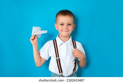 Boy Holding A Paper Airplane On A Blue Background. A Photo Of A Young Excited Schoolboy With A Happy Positive Smile Throws An Airplane Model Into The Air.