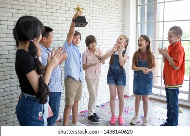 Boy Holding Trophy Cup Over His Head With Group Cute Children Student Clapping Hands Celebrating Together  In Classroom At School . Kid . Child  . Early Education .multiethnic