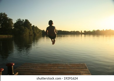 Boy Jumping Into A Lake From A Dock At Sunset In The Summer