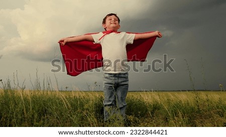 Boy kid plays superhero in red cape, childhood dream. Happy child playing superhero against sky. Little hero in red cloak looks into distance. Brave child winner dream in red raincoat plays in nature