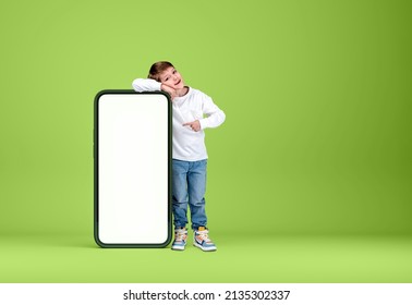 A Boy Kid Smiling, Full Length, Finger Point At Mobile Phone Mock Up Copy Space Screen, Blue Background. Concept Of Social Media And Network
