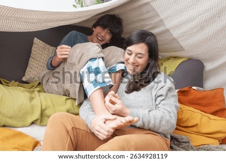 A boy laughs while sitting on a sofa under a fabric tent structure made of blankets. His mother, sitting on the floor, gently holds his feet and tickles him from below. 