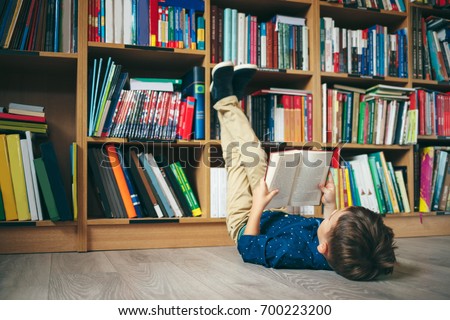 Boy laying on the floor with the feet up, reading a book against multi colored bookshelf in library. Education, Knowledge, Bookstore, Lecture. Pupil holds a book in his hands.