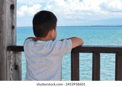 A boy leans on the railing of a wooden house terrace looking at the open sea - Powered by Shutterstock