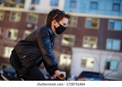 Boy In A Leather Jacket Wearing A Black Hygyenic Medical Mask With White Cross Outdoors In The Yard, Riding A Bike. View From The Side. High-rise Building On The Background. Pandemic Coronavirus 2020.