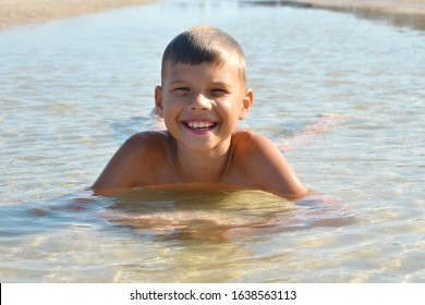 Boy Lie In Puddle On Summer. Child On Beach. Boy Lay In Dirty Water.  Happy Carefree Childhood.  Joyful Child In Muddy Puddle On Sand. Life In Village During Summer. Childhood Without Phone