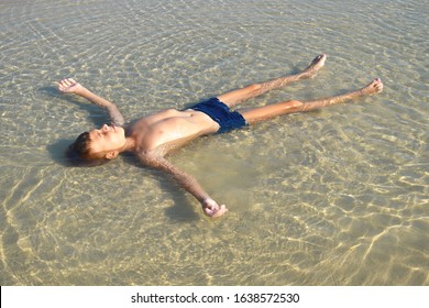 Boy Lie In Puddle On Summer. Child On Beach. Boy Lay In Dirty Water.  Happy Carefree Childhood.  Joyful Child In Muddy Puddle On Sand. Life In Village During Summer. Childhood Without Phone