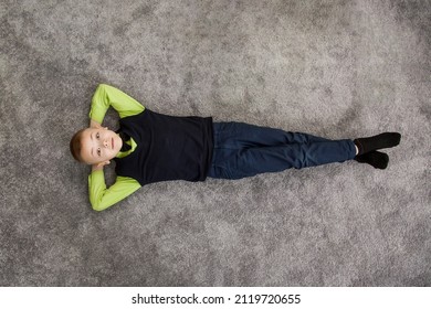 The Boy Lies On The Floor. Little Boy Lying On Carpet Floor At Home And Smiling. Child Lying Over Carpet. Happy Smiling Kid, Boy Five Years Old. Top View Of Little Boy While Lying On The Floor.