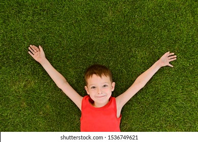 The Boy Lies On A Well-groomed Lawn. Little Boy Is Lying On The Grass. Top View Portrait Of A Young Boy Laying Down On Lawn In The Park.