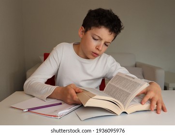 A boy  with light skin and brown hair doing homework and looking up a word in the dictionary. He is wearing a white shirt. A notebook and pencil are nearby.