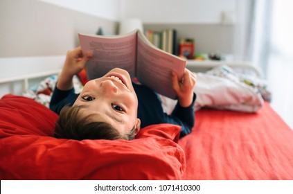 Boy Looking Back To The Camera While Reading A Book Lying On The Bed