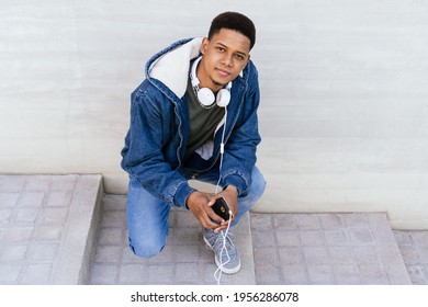 boy looking at camera using mobile phone and headphones. Afro man sitting outside on plain background. Horizontal photography, copy space.