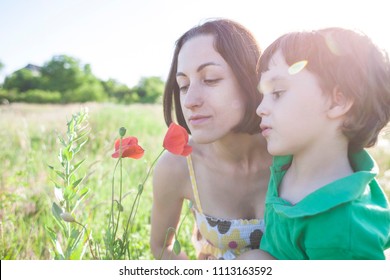 A Boy Is Looking At A Flower. A Child With Her Mother Is Sniffing Wildflowers. A Woman With A Baby Is Picking Up Poppies In A Field. Portrait Of Woman And Child In A Meadow.