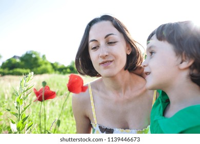 A Boy Is Looking At A Flower. A Child With Her Mother Is Sniffing Wildflowers. A Woman With A Baby Is Picking Up Poppies In A Field. Portrait Of Woman And Child In A Meadow.