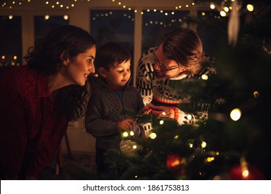 A Boy Looking At The Lights On The Christmas Tree Together With His Parents Standing By. Boy Holding A Candy Cane To Hang On Christmas Tree At Home.