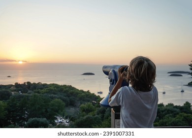  Boy looking through binocular at the islands in the sea during sunset, Vrsar, Croatia                  