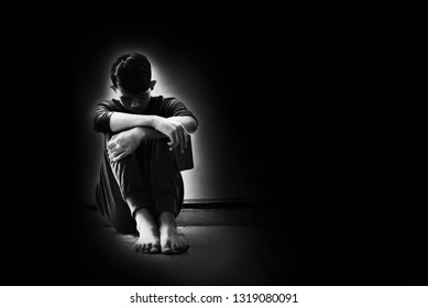 Boy with lower head and arm around knee sitting on corner of room in the dark