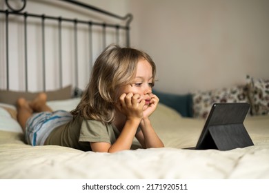 Boy lying on the bed using digital tablet computer playing games or watching cartoons at home. Child internet safety concept. Kid using electronic equipment. Parental control. Online education.