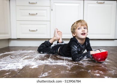 A boy lying on the kitchen floor and playing with flour. 