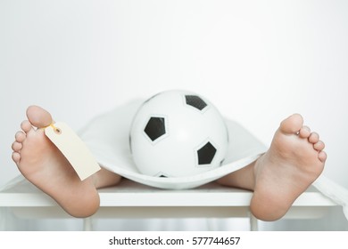 Boy Lying On A Mortuary Slab With A Soccer Ball Between His Legs On Top Of The Covering Sheet And An Identification Tag On His Toe