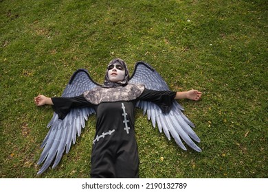 Boy In Make Up With White Face And Black Eyes, Dressed As An Angel Of Death With Wings, Lying On His Back On The Grass With A Lost Look. Concept Halloween, Autumn, Trick Or Treat, Fear, Terror.