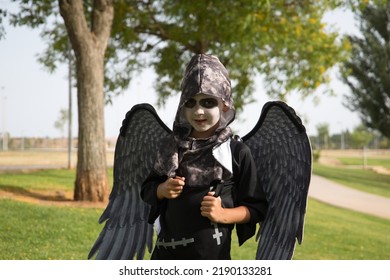 Boy In Make Up With White Face And Black Eyes, Dressed As An Angel Of Death With Wings And Haunting Look Celebrating Halloween In An Outdoor Park. Autumn Concept, Trick Or Treat, Fear, Terror.