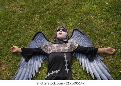 Boy In Make Up With White Face And Black Eyes, Dressed As An Angel Of Death With Wings, Lying On His Back On The Grass With A Lost Look. Concept Halloween, Autumn, Trick Or Treat, Fear, Terror.