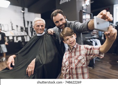 The boy makes selfie on a smartphone with two older men in barbershop, where they came to get a haircut. One of the men is sitting in a hairdresser's armchair. They smile and have a good mood. - Powered by Shutterstock