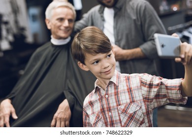 The boy makes selfie on a smartphone with two older men in barbershop, where they came to get a haircut. One of the men is sitting in a hairdresser's armchair. They smile and have a good mood. - Powered by Shutterstock