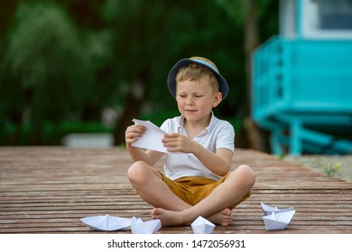 Boy Making Paper Boat On Wooden Pier In The Beach. Vacation Concept. Paper Ship In Child Hands. Little School Boy In White T-shirt And Hat On Sunny Day.