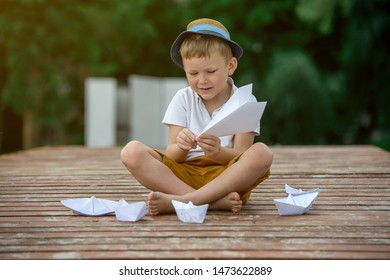 Boy Making Paper Boat On Wooden Pier In The Beach. Vacation Concept. Paper Ship In Child Hands. Little School Boy In White T-shirt And Hat On Sunny Day.