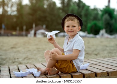 Boy Making Paper Boat On Wooden Pier In The Beach. Vacation Concept. Paper Ship In Child Hands. Little School Boy In White T-shirt And Hat On Sunny Day.