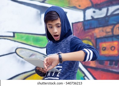 Boy, Man In Blue Hoody Holding Tablet Pad Running Late To School Looking To His Wrist Watch Isolated Graffiti Wall Background. Outdoors, Lifestyle Horizontal Image