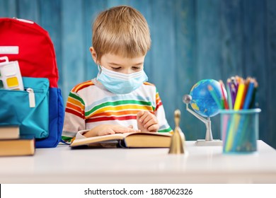 Boy In Medical Face Protection Mask Indoors On Blue Background. Sick Child Sitting At The Table And Reading A Book. Coronavirus Pandemic.