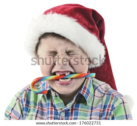 A boy with missing teeth wearing a Santa hat biting on a candy cane.