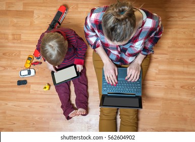 Boy and mother using tablet and laptop while playing with toy cars on a carpet at home. Modern family. Tablet pc hero header image. Boy using digital tablet while sitting on wooden floor. Work at home