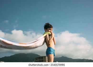 A Boy On The Beach With A Towel That Flutters In The Wind Like A Superman Cape. Happy Child In Summer On The Background Of Mountains. High Quality Photo