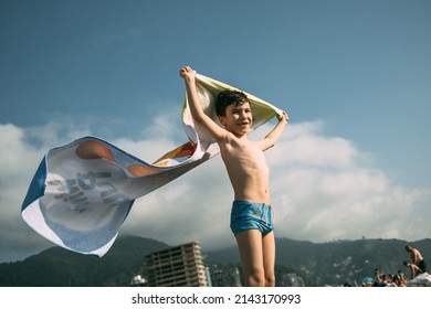 A Boy On The Beach With A Towel That Flutters In The Wind Like A Superman Cape. Happy Child In Summer On The Background Of Mountains. High Quality Photo