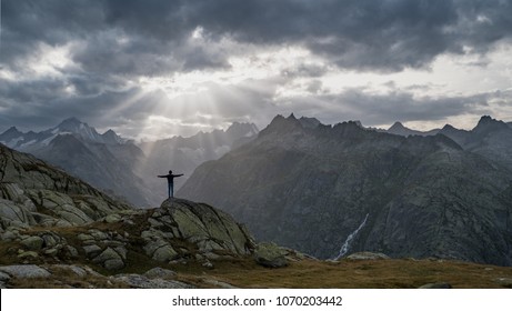 A boy on a hiking trip enjoys an incredible view of sun rays breaking through thick dark clouds over a valley, posing in "Jesus" or christian cross pose with half raised arms