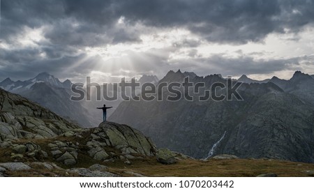 A boy on a hiking trip enjoys an incredible view of sun rays breaking through thick dark clouds over a valley, posing in 