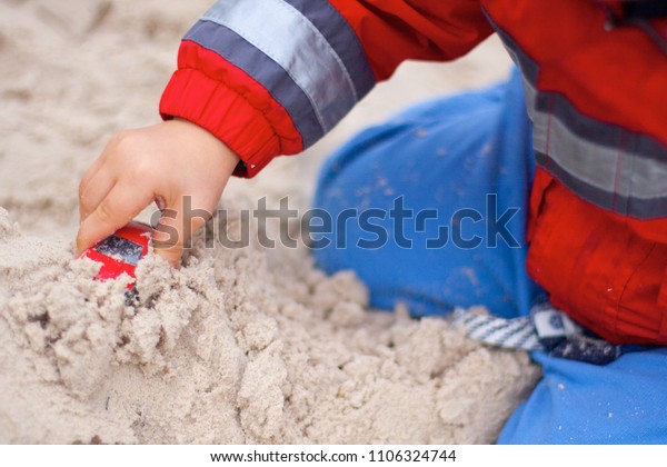 A boy on the playground playing in\
a sandpit with a red toy car. A view of legs and\
arms.