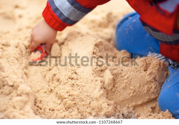 A boy on the playground playing in\
a sandpit with a red toy car. A view of legs and\
arms.