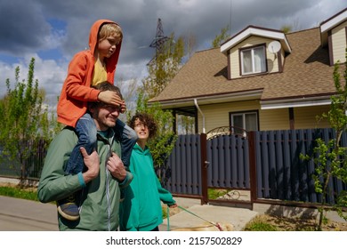 A boy in an orange hoodie is sitting on his father's neck, his mother is walking next to him. They are walking down the street near the house in the village